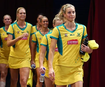 Australian netball team players in yellow uniforms walking on court, holding towels and water bottles.