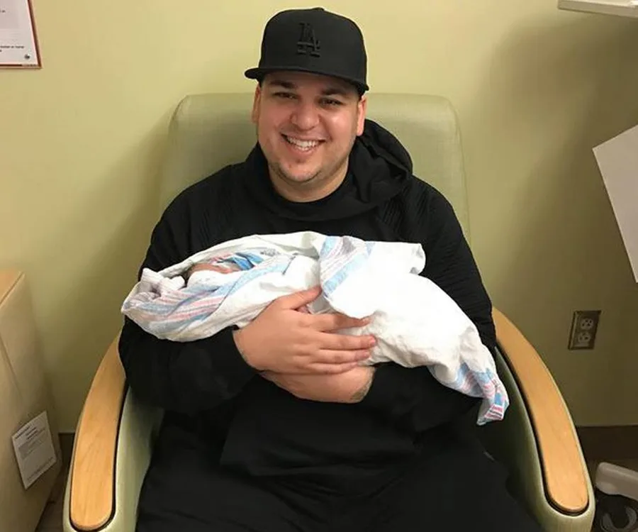 Person in black holding a newborn wrapped in a hospital blanket, seated in a chair, smiling.