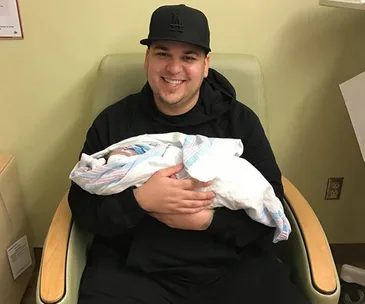 Person in black holding a newborn wrapped in a hospital blanket, seated in a chair, smiling.