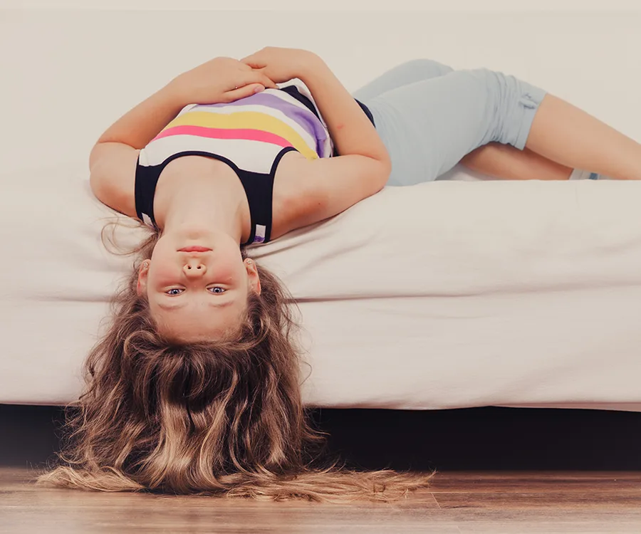 Girl lying upside down on a bed, wearing a striped tank top and shorts, with long hair hanging off the edge.