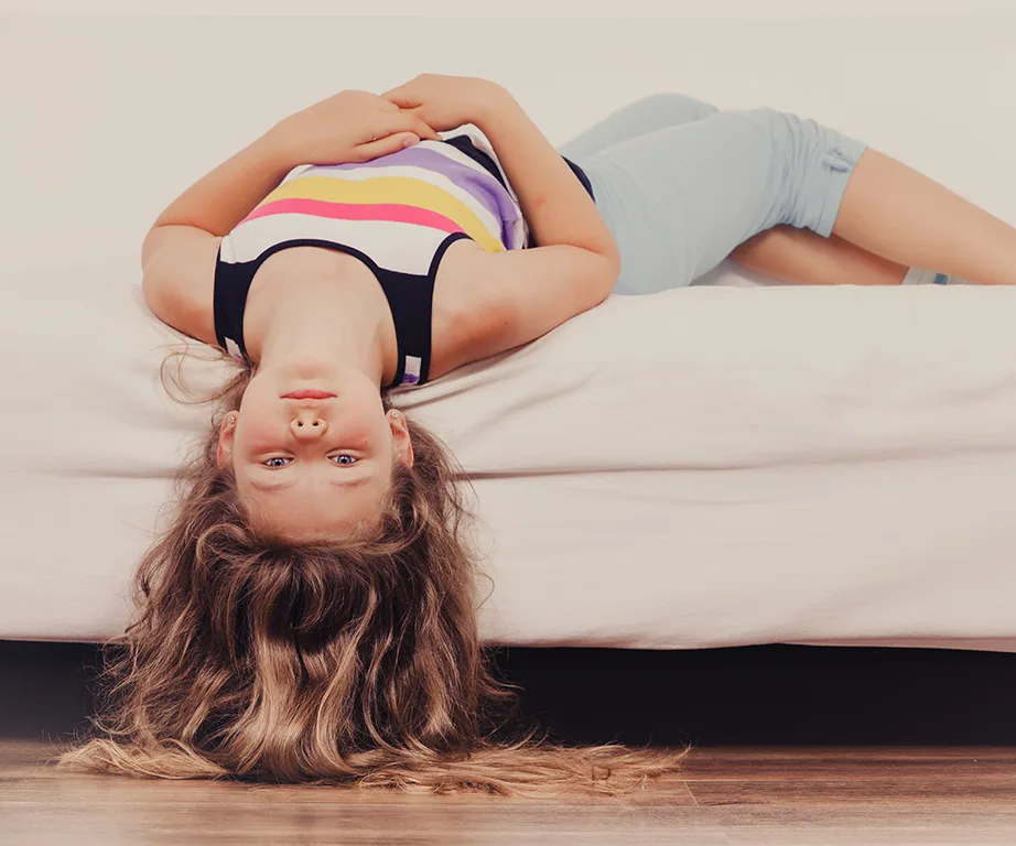 Girl lying upside down on a bed, wearing a striped tank top and shorts, with long hair hanging off the edge.
