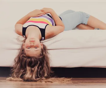 Girl lying upside down on a bed, wearing a striped tank top and shorts, with long hair hanging off the edge.