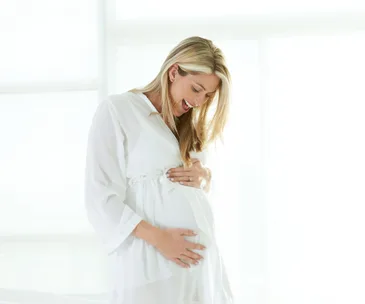 Pregnant woman in white robe lovingly holding her belly, smiling, with a bright window in the background.