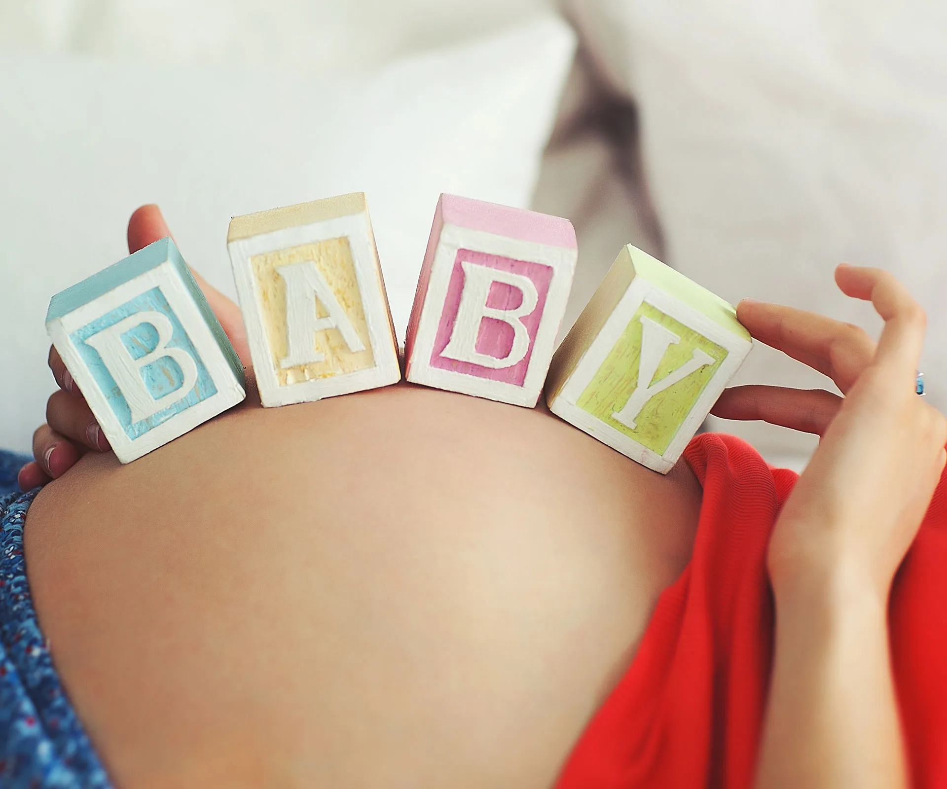 Pregnant belly with "BABY" spelled on wooden blocks, woman in red shirt, close-up.