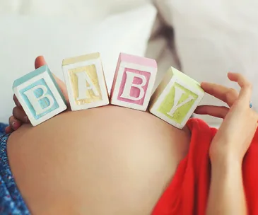 Pregnant belly with "BABY" spelled on wooden blocks, woman in red shirt, close-up.