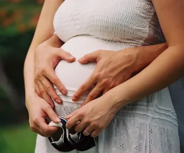 Pregnant woman at 38 weeks in white dress, with hands making heart shape on belly, holding baby shoes.