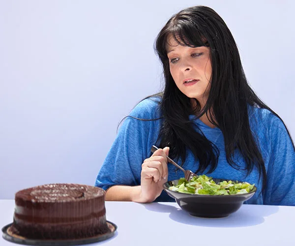 Woman in blue shirt, looking at chocolate cake on left, eating salad, expression thoughtful.