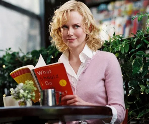 A woman with curly blonde hair, wearing a pink sweater, sits at a cafe table reading "What Should I Do with My Life?"