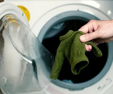 Hand holding a small green sweater near an open washing machine door.