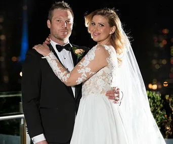 A bride in a white gown and a groom in a tuxedo embrace at an outdoor nighttime setting.