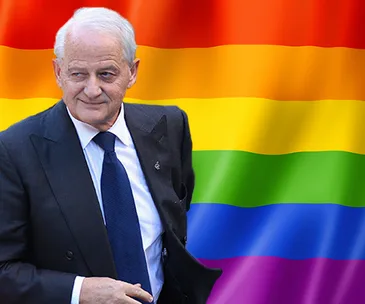 A man in a dark suit stands in front of a rainbow flag background.