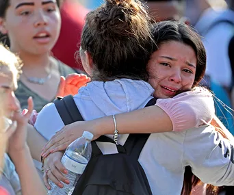 Two people hugging, one visibly upset, holding a water bottle, with others in the background, likely in a comforting moment.