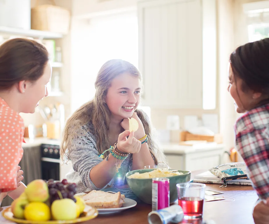 Teenagers gathered in a kitchen, smiling and talking while snacking on chips and fruit at a table.
