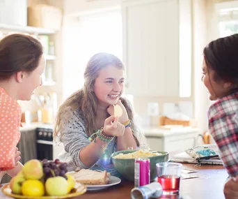 Teenagers gathered in a kitchen, smiling and talking while snacking on chips and fruit at a table.