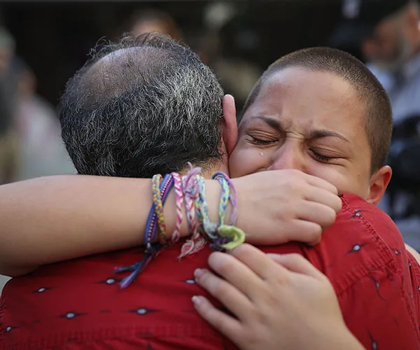 A person with short hair and emotional expression hugs another tightly, wearing colorful bracelets, conveying comfort and support.