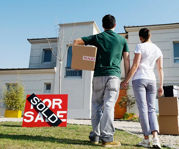 Couple holding a box stands outside a newly purchased house with "Sold" sign.