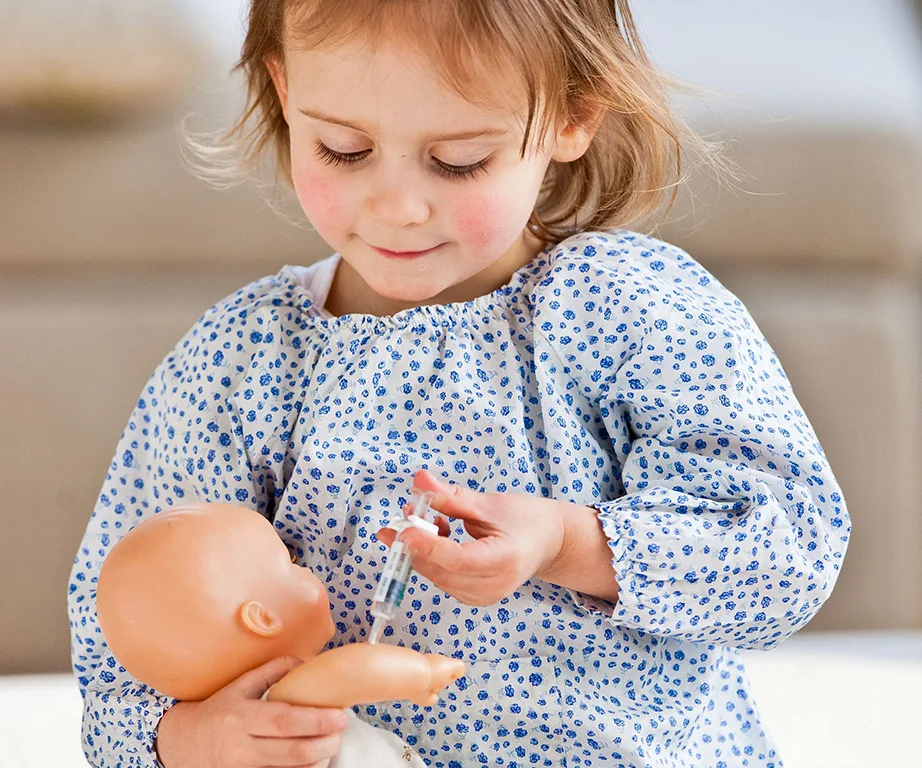Young child in blue floral shirt giving a toy syringe to a doll.