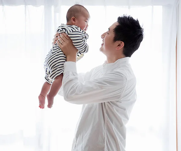 A man in a white shirt holds a baby in a striped outfit up towards a window, bright light in the background.