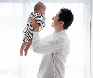 A man in a white shirt holds a baby in a striped outfit up towards a window, bright light in the background.
