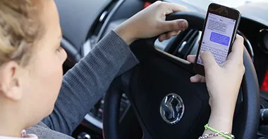 A person texting on a smartphone while driving, with hands on the wheel of a Volkswagen car.