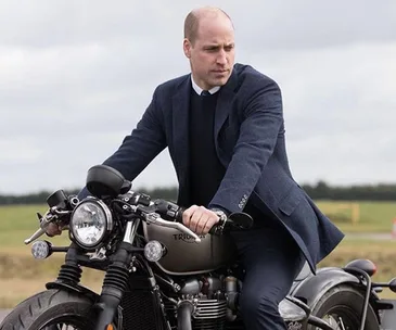Man in a suit sits on a Triumph motorcycle outdoors, looking thoughtful.