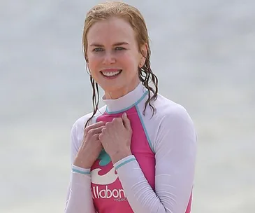 Woman smiling in a pink and white wetsuit on a beach, with the ocean in the background.