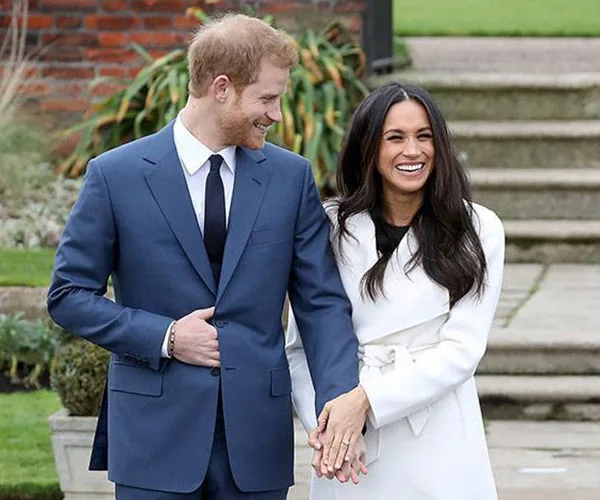 Prince Harry and Meghan Markle laughing and holding hands outdoors, dressed in a blue suit and white coat respectively.