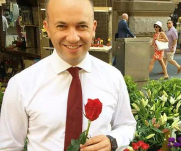 A man in a white shirt and red tie holds a red rose, standing in front of a flower stall on a city street.