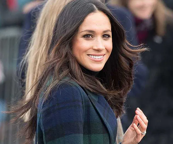 Smiling woman with long hair in a plaid coat, facing sideways outdoors.