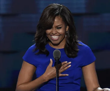 Michelle Obama smiling, wearing a blue dress, with one hand on her chest, standing in front of a microphone.