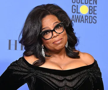 Oprah Winfrey at the Golden Globe Awards, wearing a black dress and glasses, with a blue background featuring event logos.
