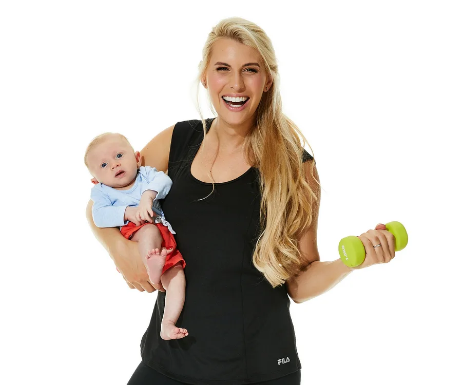 Woman holding a baby and a green dumbbell, smiling against a white background.