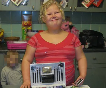 Child in a red striped shirt smiling, standing in a kitchen, with open makeup set on table and pink toy horse in background.