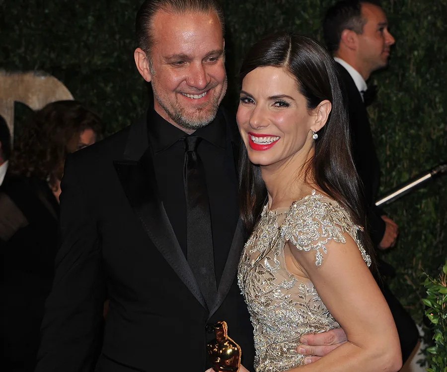 A smiling woman in an ornate dress holds an Oscar statue, standing close to a man in a black suit.