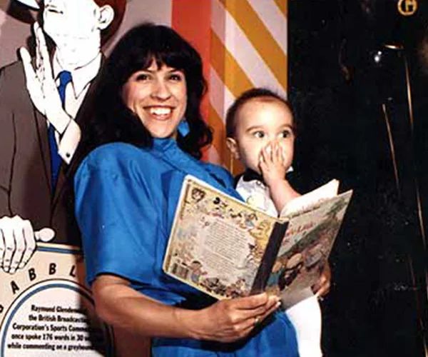 Woman in blue holding a child and a book, smiling, with a cartoon figure in the background.