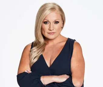 Blonde woman with arms crossed, gazing confidently; wearing a dark blue top against a plain background.