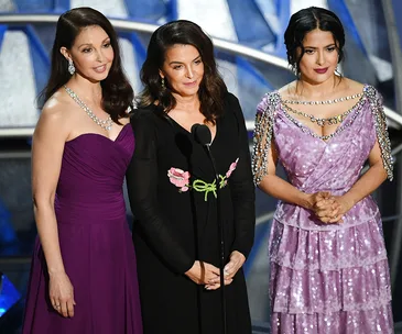 Three women stand together on stage at the 2018 Oscars, dressed elegantly in evening gowns.