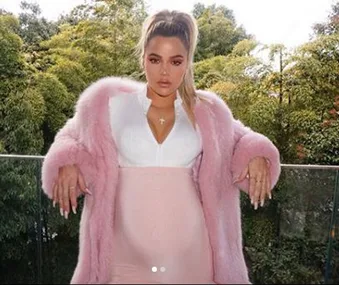 Khloe Kardashian in a pink coat and dress, standing outdoors with greenery in the background.