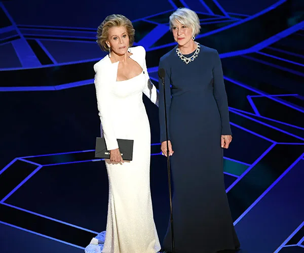 Jane Fonda and Helen Mirren presenting at the Oscars in elegant gowns, standing on a geometric-patterned stage.