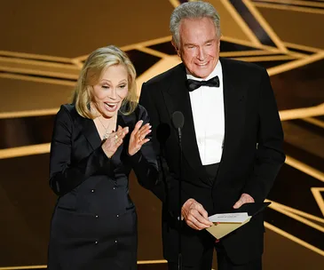 Actors presenting an award at the Oscars, one holds an envelope, both are smiling on stage.