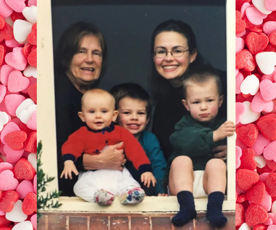 A woman and a younger woman smiling with three children on a window ledge against a backdrop of colorful hearts.