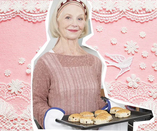 Elderly woman in a headband smiling, holding a tray of baked goods against a pink, lace-patterned background.