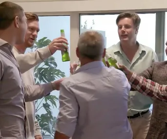 A group of men inside a room raising beer bottles in a toast gesture, standing around a table with a window in the background.