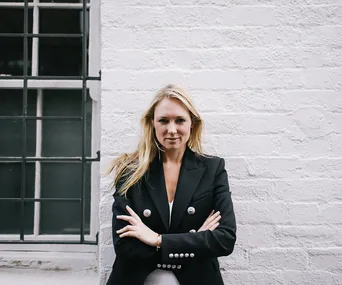 Person in a black blazer standing against a white brick wall, arms crossed, looking confidently at the camera.
