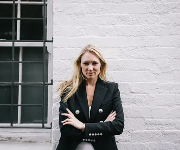 Person in a black blazer standing against a white brick wall, arms crossed, looking confidently at the camera.
