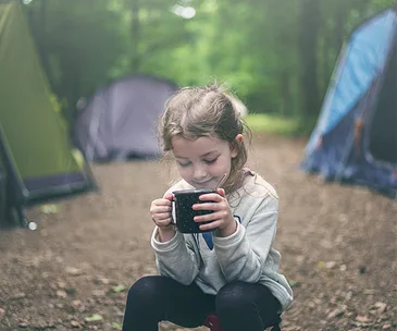 Young girl drinking from a mug while sitting outdoors at a campsite with tents in the background.