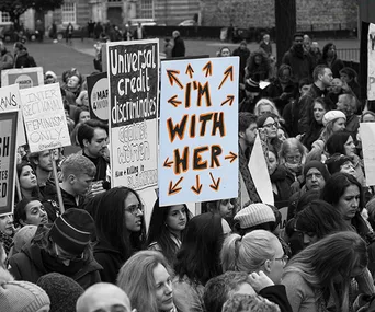 Crowd at a rally with "I'm With Her" and "Universal Credit Discriminates" signs visible.