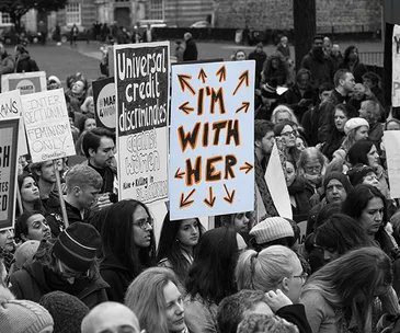 Crowd at a rally with "I'm With Her" and "Universal Credit Discriminates" signs visible.