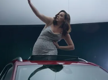 Woman in silver dress poses dramatically on red car roof.