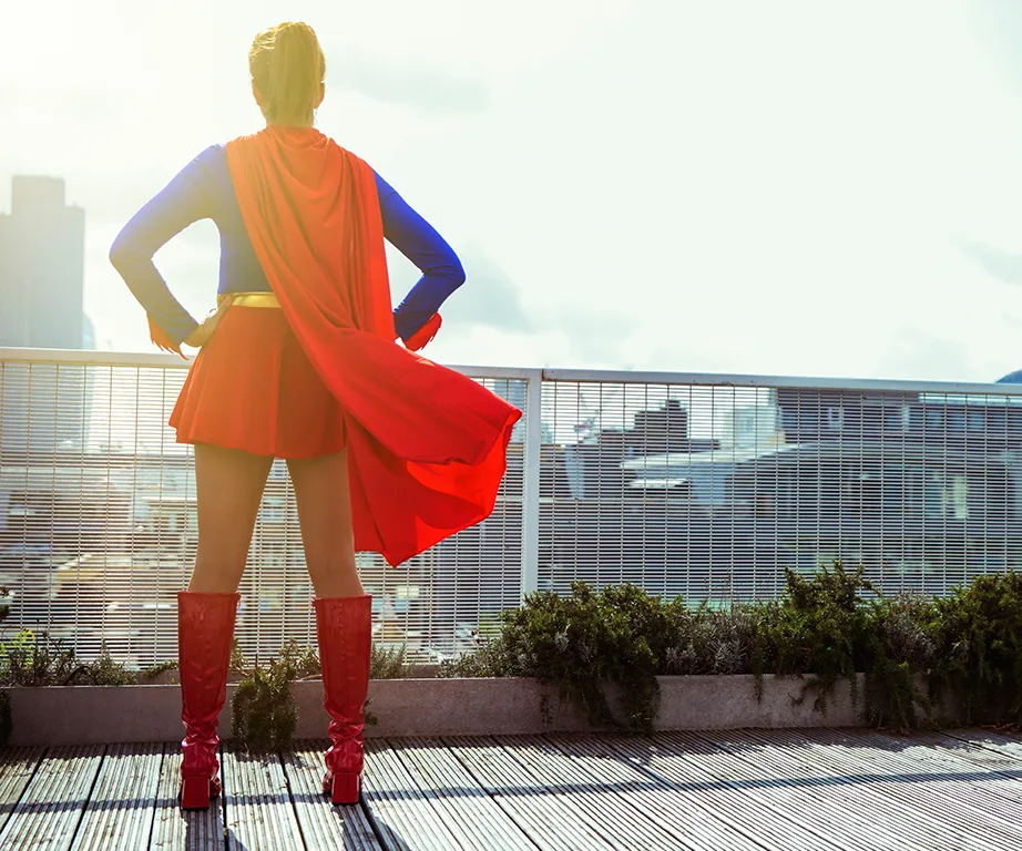 A woman in a superhero costume stands on a rooftop, gazing at the city skyline, with a red cape flowing behind her.